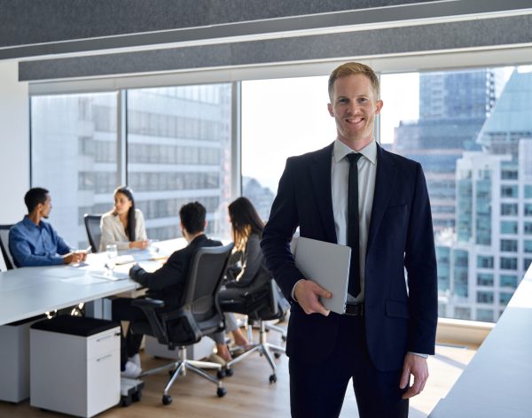 Smiling confident young business man manager, businessman corporate leader, male ceo boss wearing suit standing in office on team workers background working in board meeting room office, portrait.