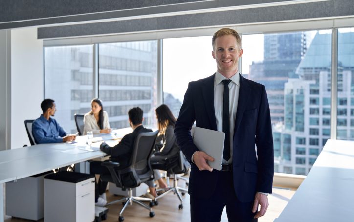 Smiling confident young business man manager, businessman corporate leader, male ceo boss wearing suit standing in office on team workers background working in board meeting room office, portrait.