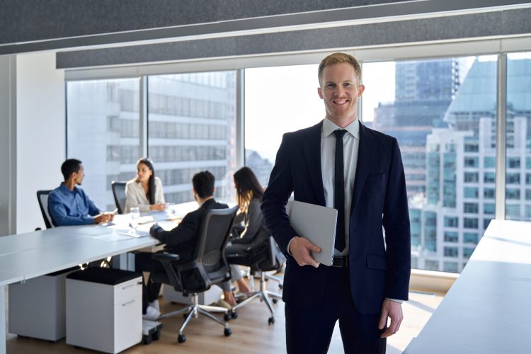 Smiling confident young business man manager, businessman corporate leader, male ceo boss wearing suit standing in office on team workers background working in board meeting room office, portrait.