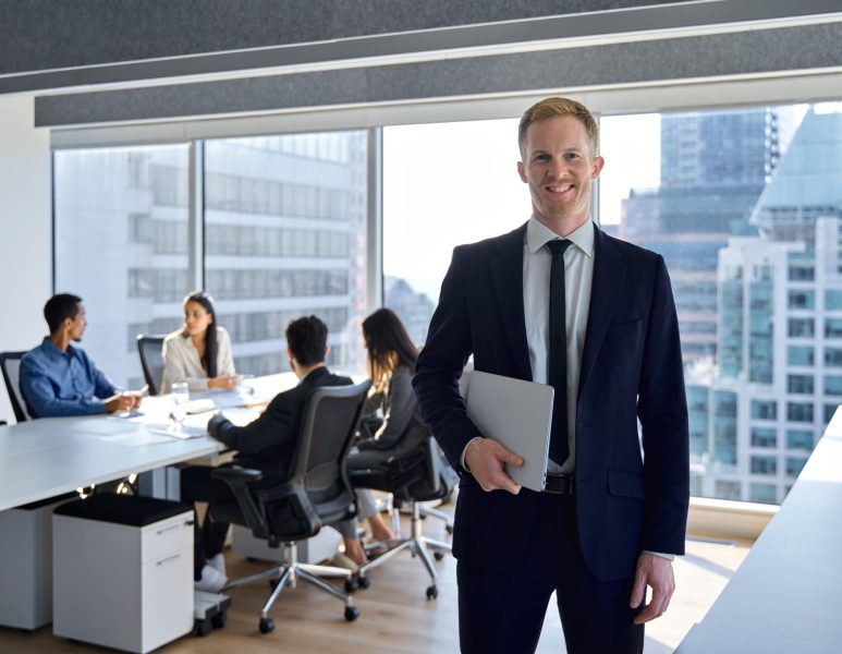 Smiling confident young business man manager, businessman corporate leader, male ceo boss wearing suit standing in office on team workers background working in board meeting room office, portrait.